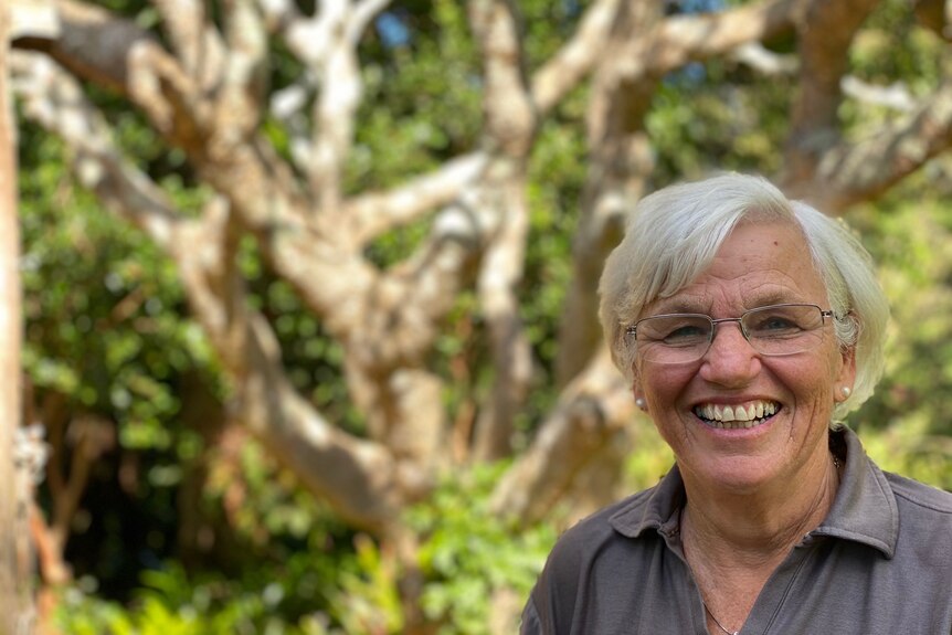 A woman with gray hair smiles while standing in front of a frangipani tree devoid of flowers and leaves