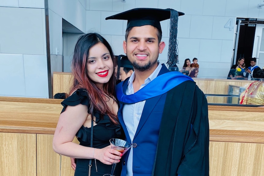 A young man wears a graduation cap and gown next and stands next to a young lady wearing a black dress