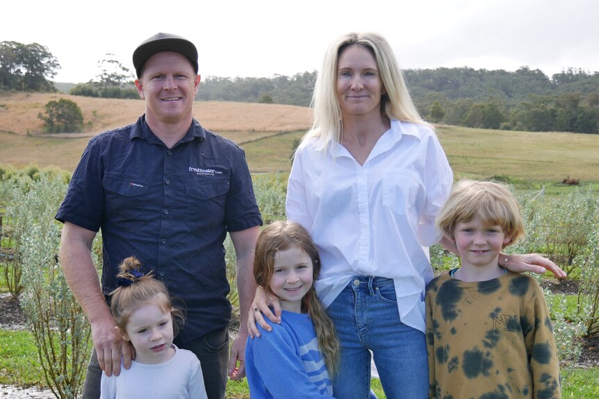 A smiling couple with their three children standing in front of salt bush plants, man wears cap, blue shirt, woman white shirt.