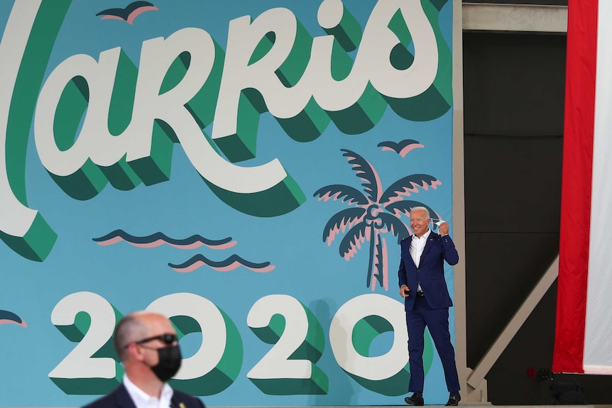 Joe Biden walking past a colourful sign while removing his face mask