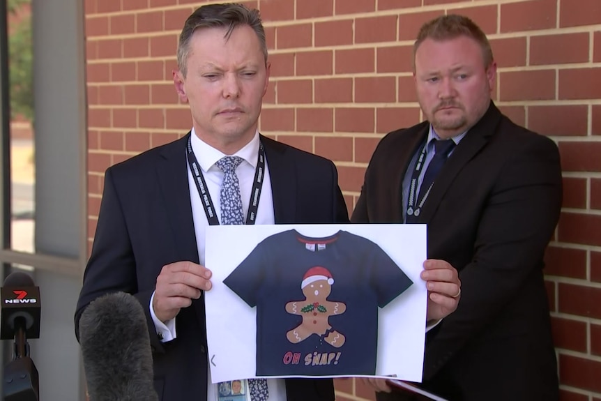 A wide shot of a police officer holding a picture of a pyjama top with another officer standing next to a wall behind him.,