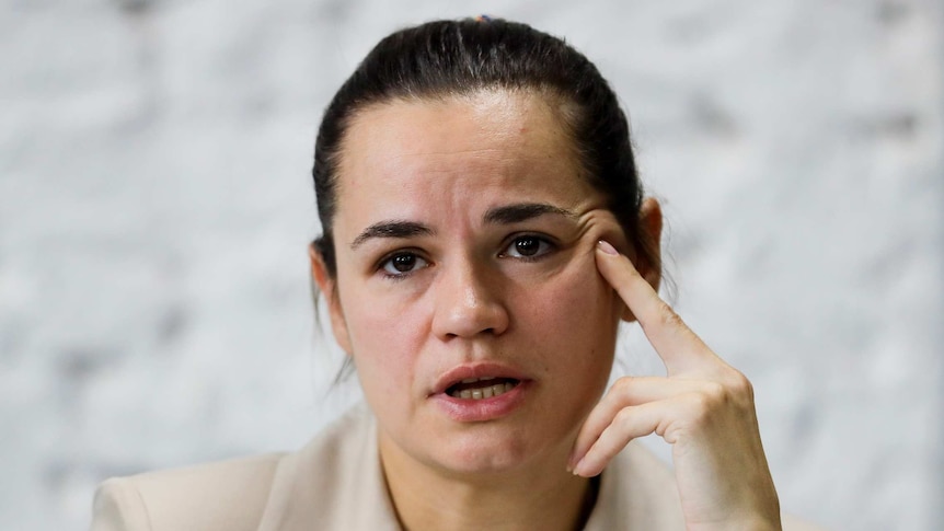 A woman with dark hair rests her left middle finger on her left cheek as she speaks