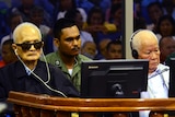 Two elderly Cambodian men, one wearing sunglasses, sit in a courtroom with headphones on.