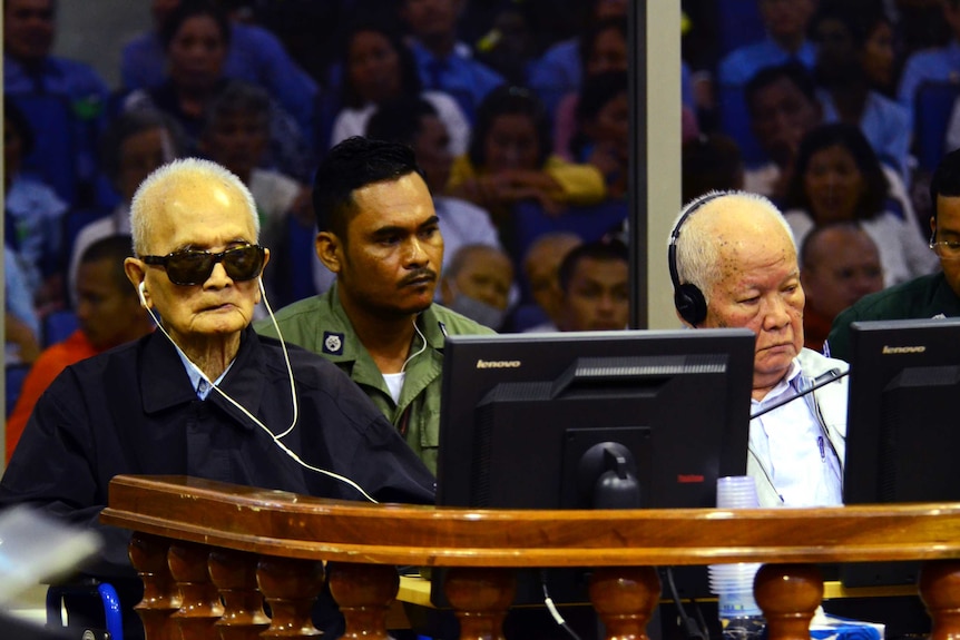 Two elderly Cambodian men, one wearing sunglasses, sit in a courtroom with headphones on.