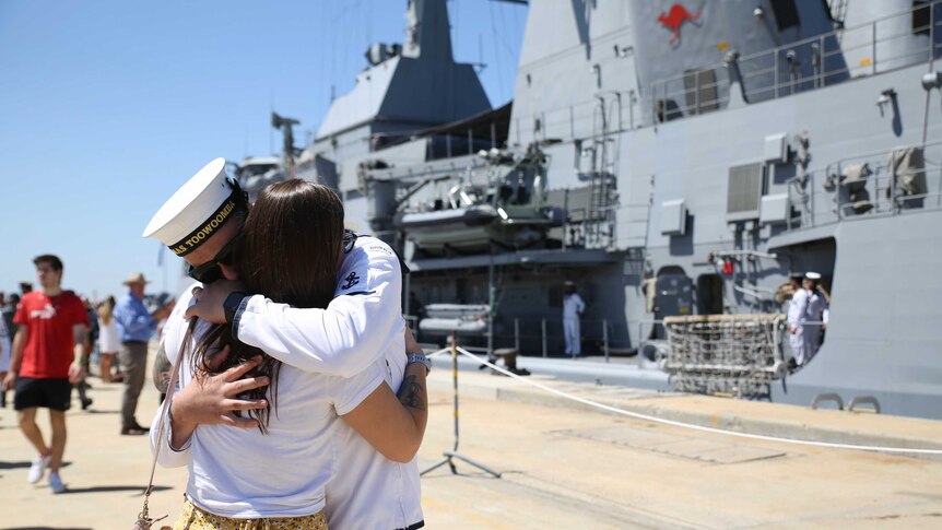 Two people embrace in front of a huge navy ship