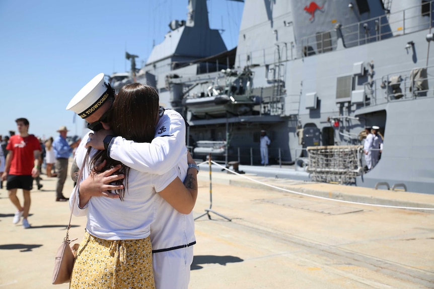 Two people embrace in front of a huge navy ship