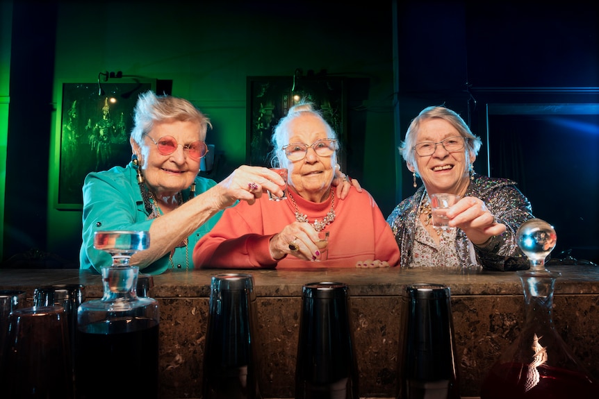 Three smiling elderly women, dressed up, sitting in a dark bar, having shots.