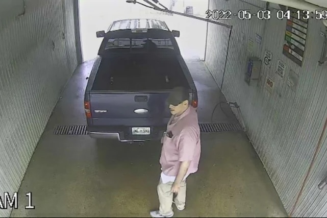 Un grand homme vêtu d'une chemise rose et d'une casquette de baseball foncée se promène dans un ute sombre dans une baie de lavage de voiture