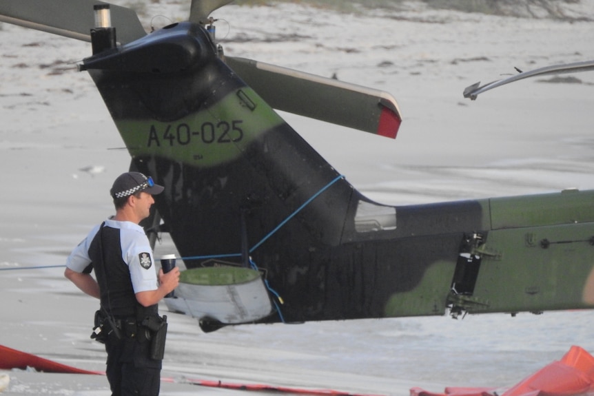 A police officer holding a cup of coffee stands beside a miltiary helicopter on a beach.