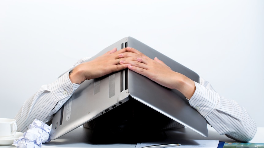 Person sits buried by lap-top held over top of their head, which rests on a table with a screwed up piece of paper. 