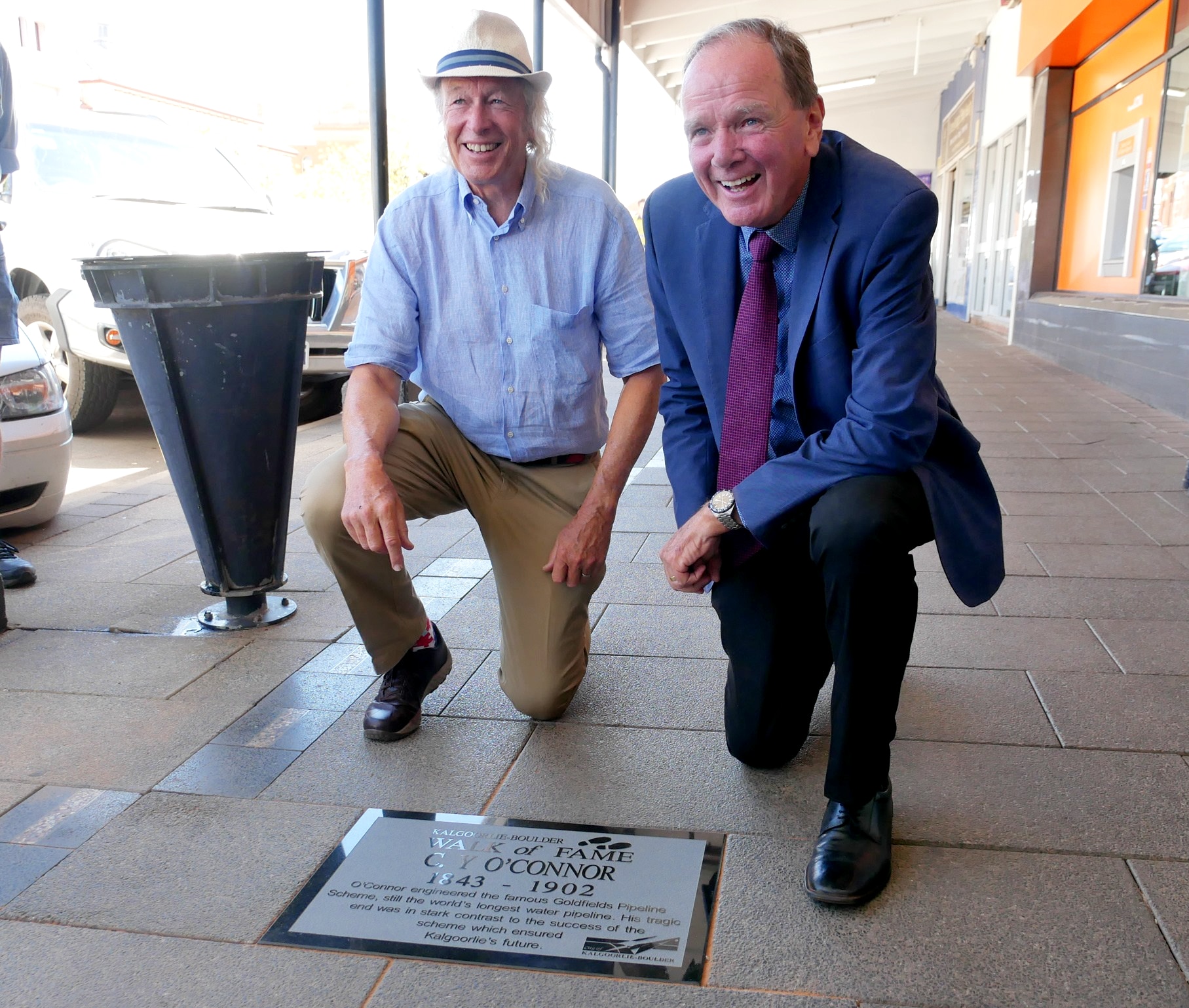 Two men kneeling over a plaque on a footpath. 