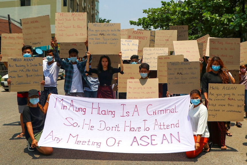 A crowd of people hold signs in the middle of a street on a clear day, each carrying words criticising Min Aung Hlaing. 