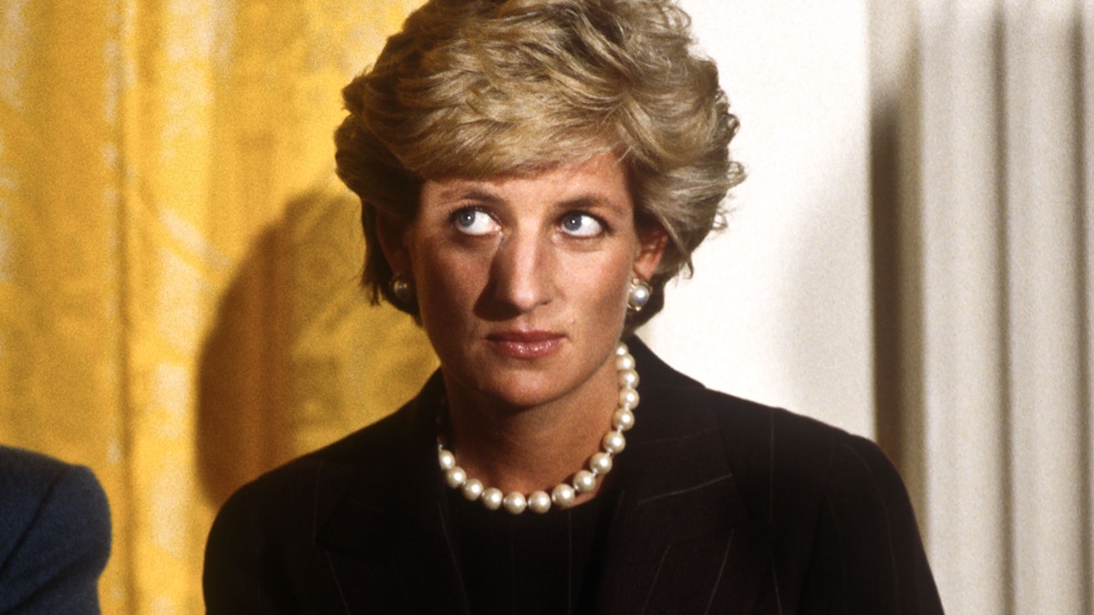 Princess Diana with short blonde hair wears a black suit and white pearl necklace with matching earrings and looks off camera.