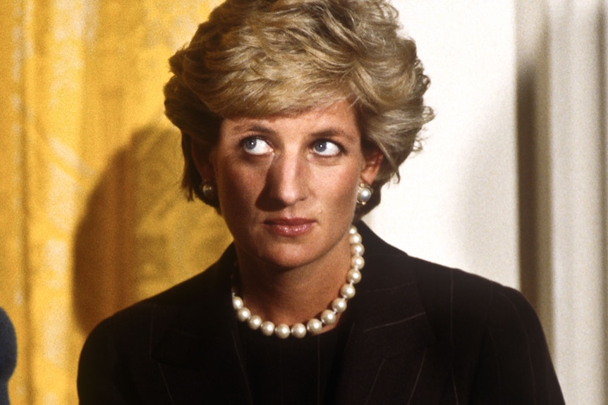 Princess Diana with short blonde hair wears a black suit and white pearl necklace with matching earrings and looks off camera.