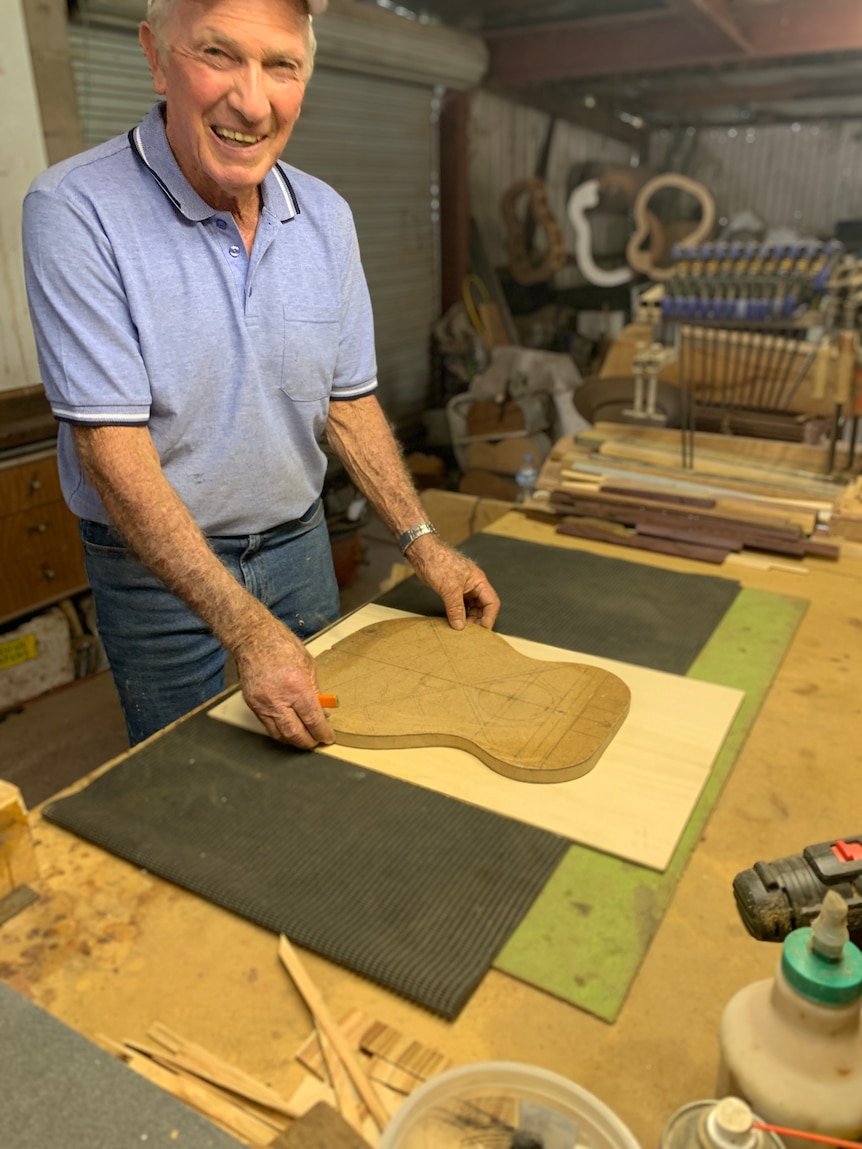 A man in a blue t-shirt smiles while standing at a workshop table.