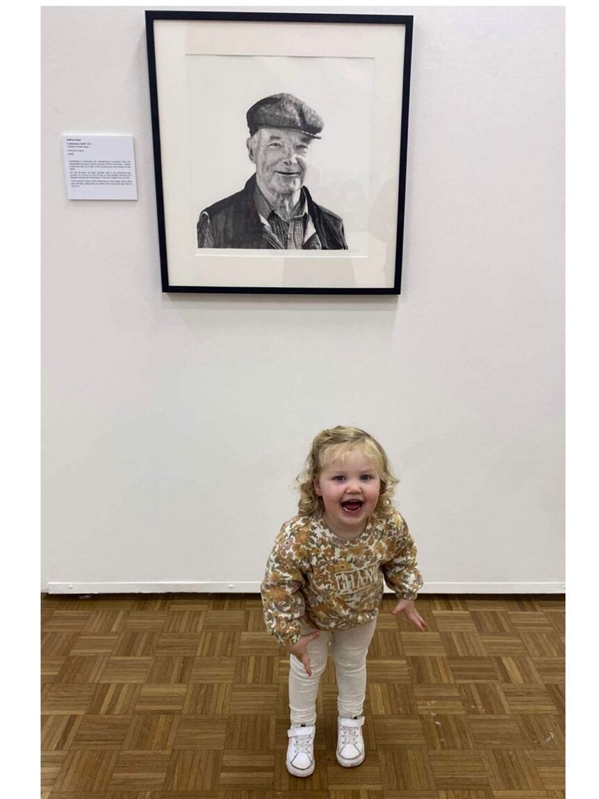 A toddler grins wildly standing underneath a portrait of her great grandfather
