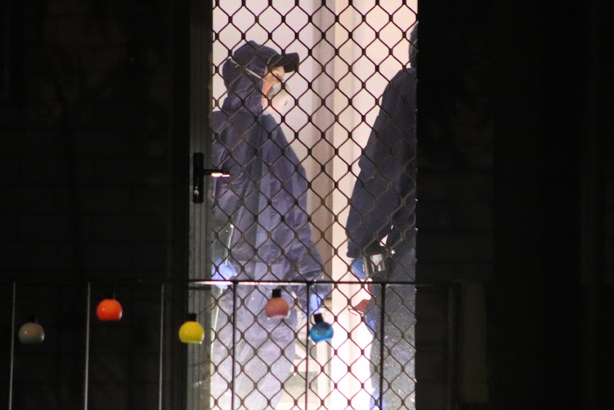 A police forensic officer in a jump suit and face mask stands inside the door of a house at night.