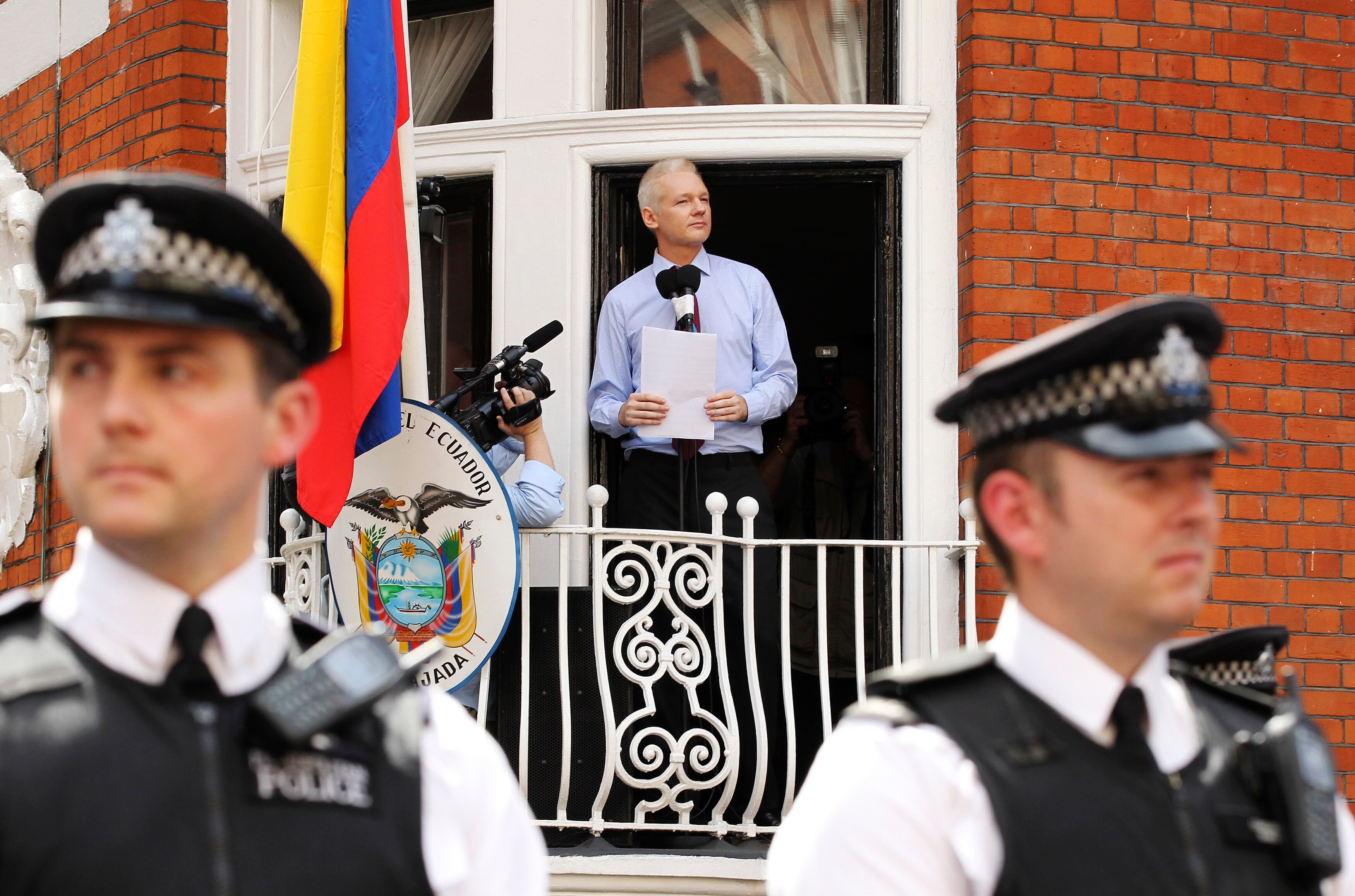 Two police officers stand guard outside the embassy, where Julian is standing on a balcony giving a speech.
