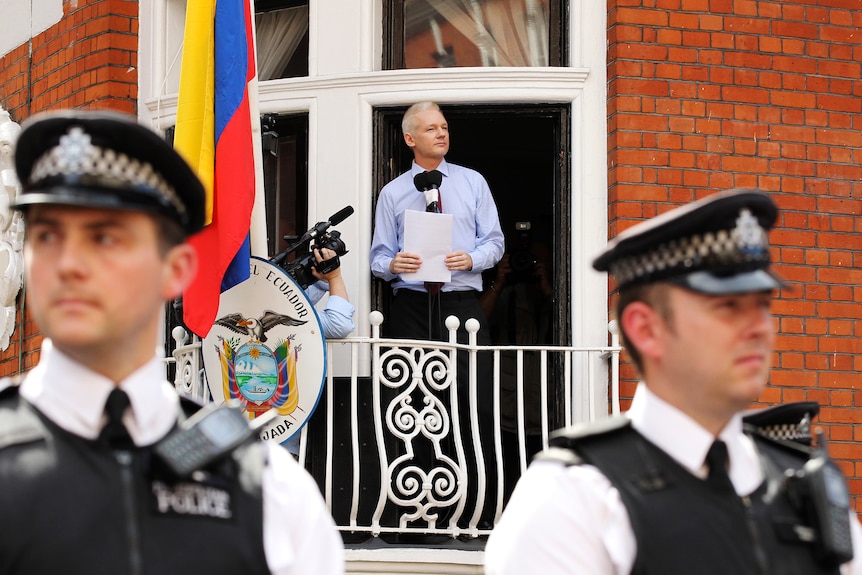 Two police officers stand guard outside the embassy, where Julian is standing on a balcony giving a speech.
