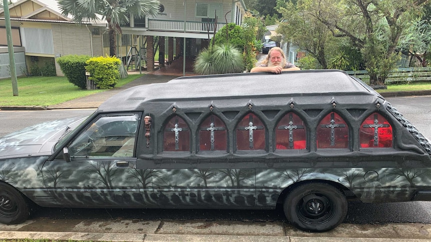 Karl Claydon leans on his converted hearse with gothic windows, a skeleton and a cemetery scene on the side parked on the street
