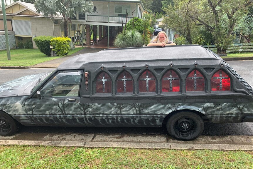 Karl Claydon leans on his converted hearse with gothic windows, a skeleton and a cemetery scene on the side parked on the street