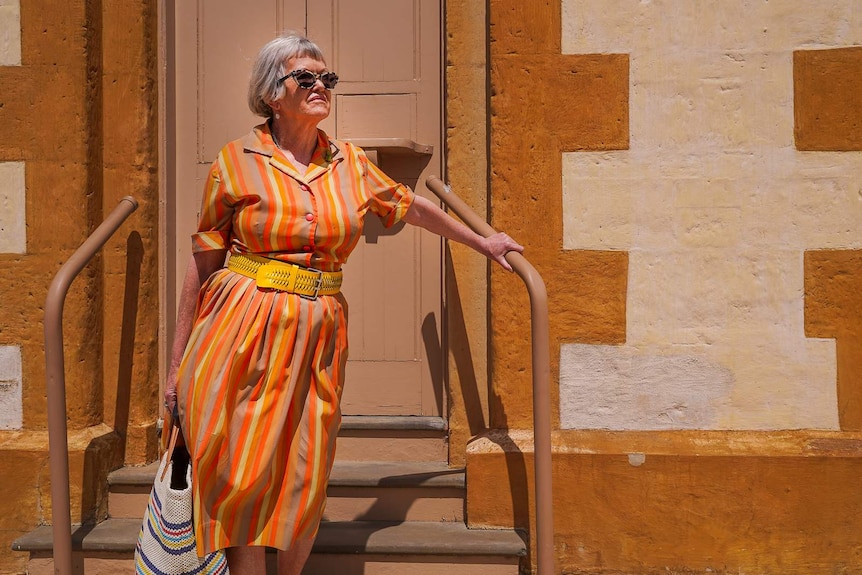 A woman in an orange and yellow striped frock and bold sunglasses stands on the front steps of an old stone building.