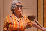 A woman in an orange and yellow striped frock and bold sunglasses stands on the front steps of an old stone building.