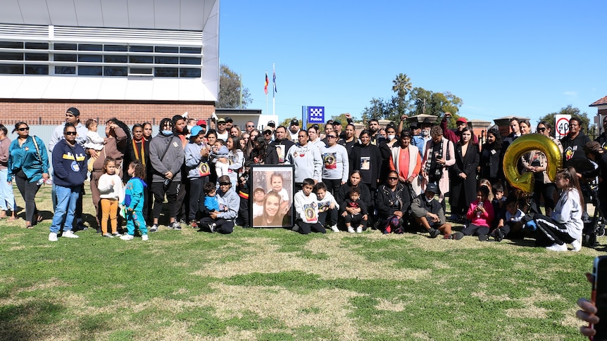 Dozens of people standing on grass around a framed photograph. 