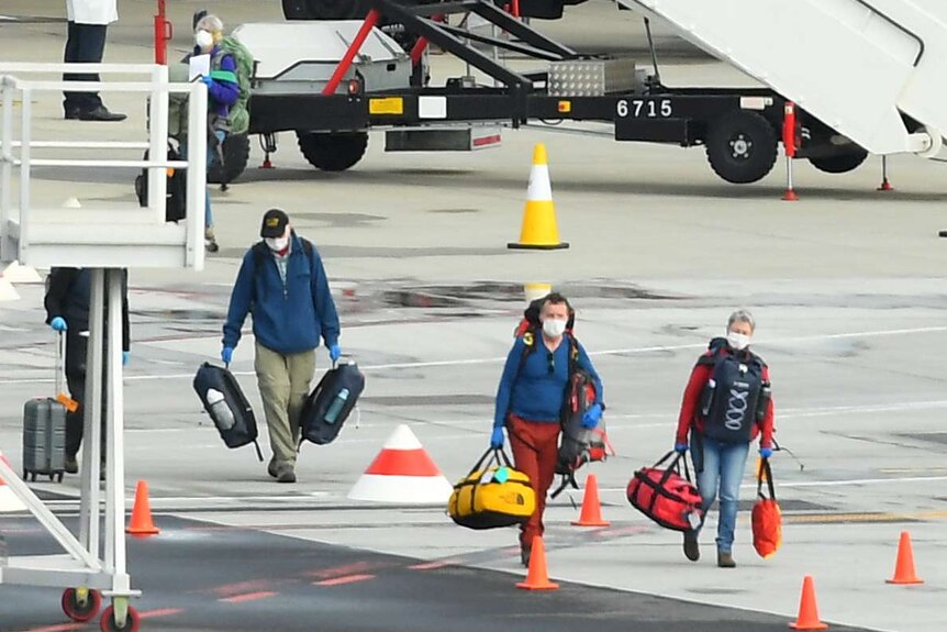 Masked passengers walk across the tarmac of Melbourne Airport with bags in their hands.