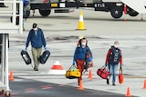 Masked passengers walk across the tarmac of Melbourne Airport with bags in their hands.
