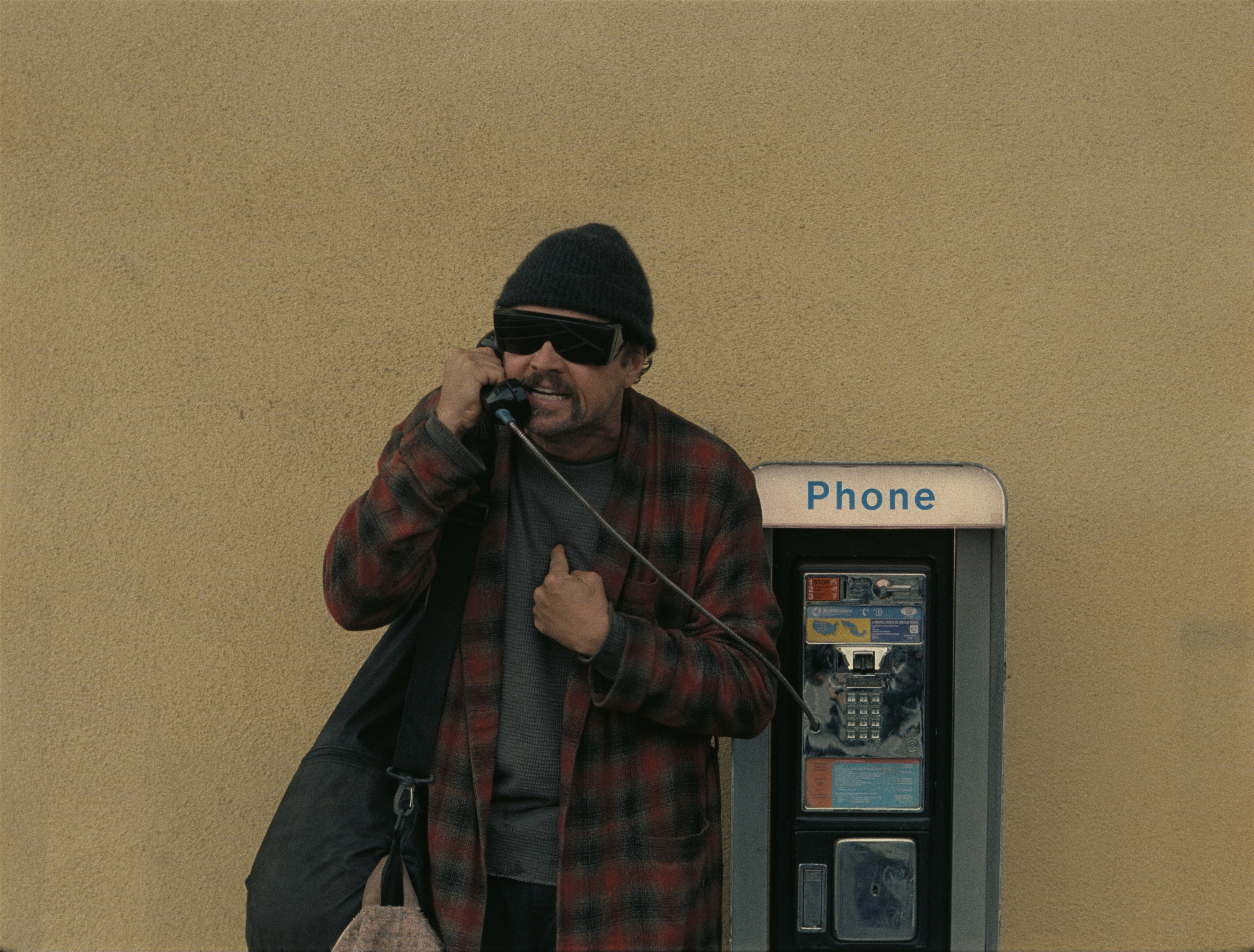Leo DiCaprio in a beanie and big, dark glasses talks aggressively into a payphone