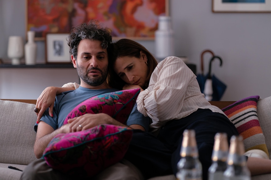 Arian Moayed, and Iranian man with curly dark hair and stubble, and Michaela Watkins, a brunette white woman, snuggle on a couch