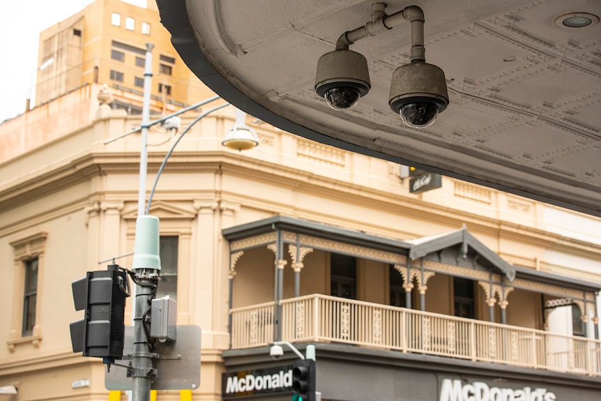 Deux caméras de vidéosurveillance sont suspendues à une véranda avec des bâtiments en arrière-plan. 