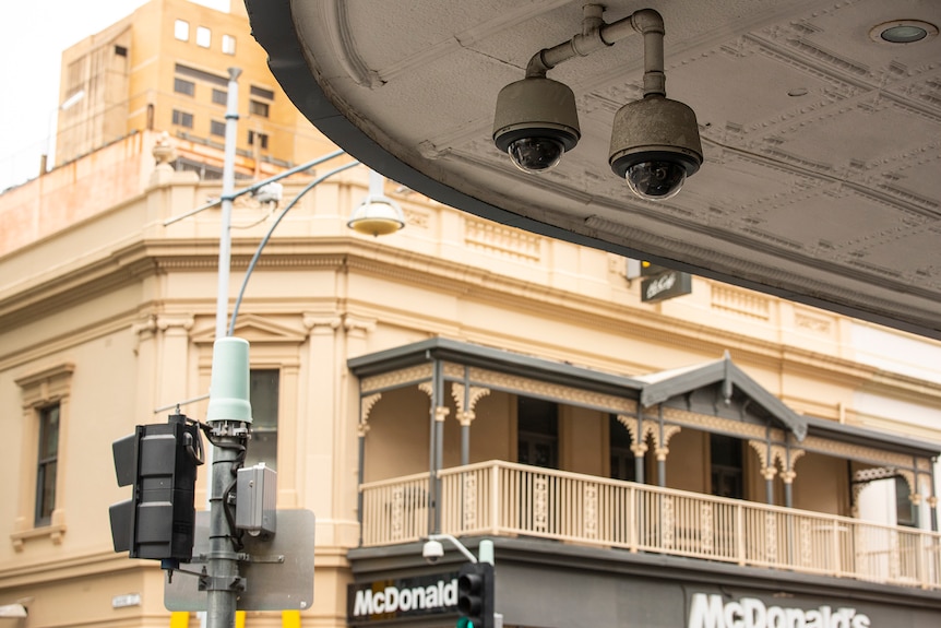 Two CCTV cameras hang from a veranda with buildings in the background. 