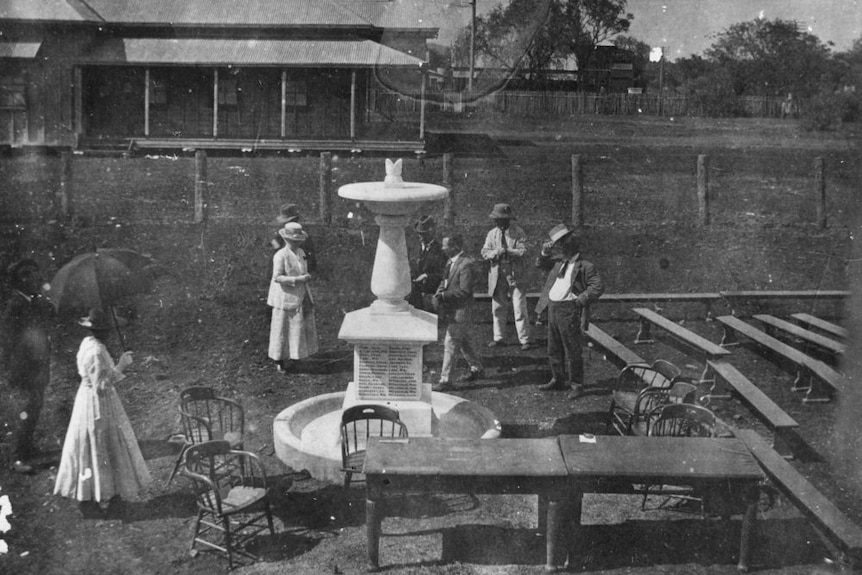 A black and white image of a small war memorial with some people standing nearby. 