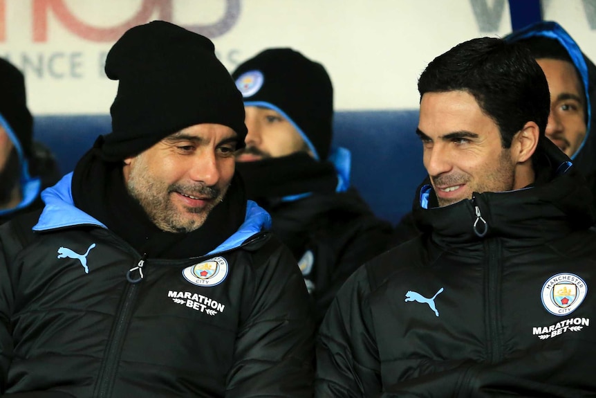 Pep Guardiola smiles sitting next to Mikel Arteta in the Manchester City dugout