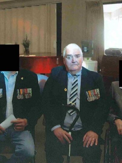 An elderly man with war medals on his jacket in a nursing home with other unidentified residents.