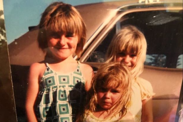 An old photograph of three young girls leaning against a car in the afternoon light grinning.