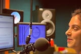 A photo of Nas Campanella sitting behind a radio microphone, surrounded by computers and speakers.