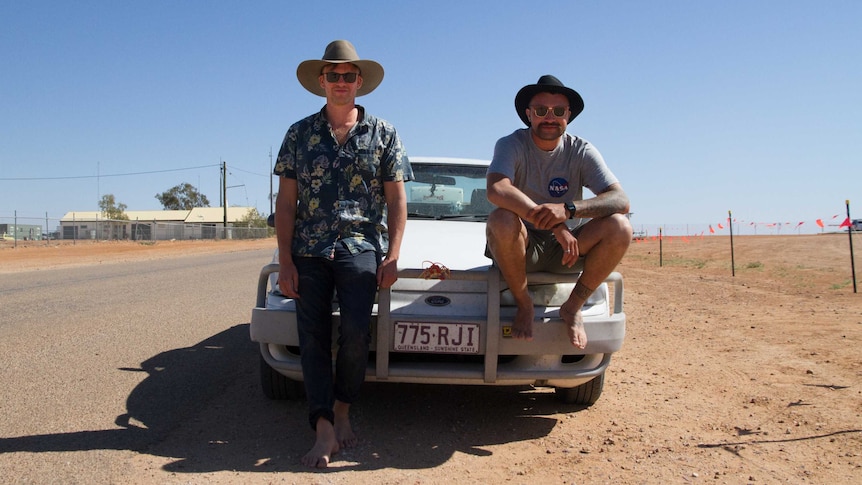 Two men with wide-brimmed hats lean against and sit on the bullbar and bonnet of their car. There is red dirt by the roadside.