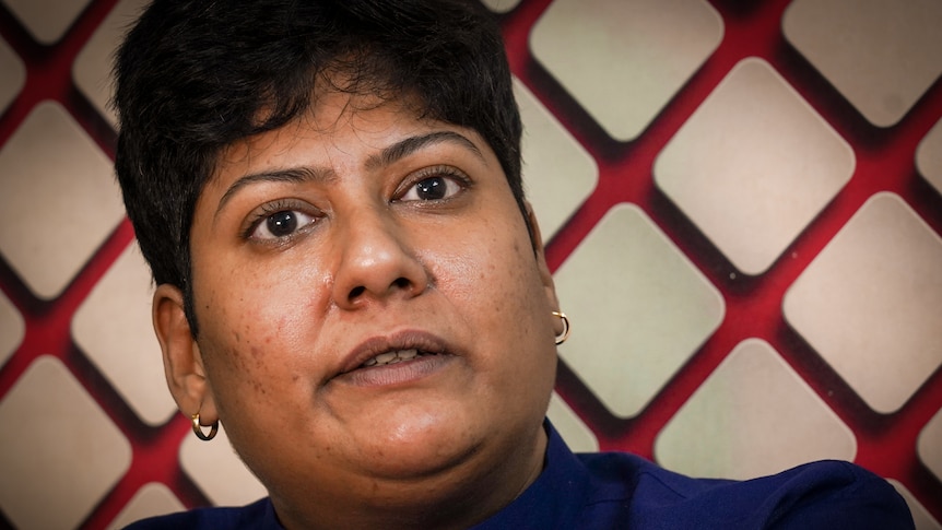 A woman with cropped black hair sits against a red and white decorative wall with a thoughtful expression 