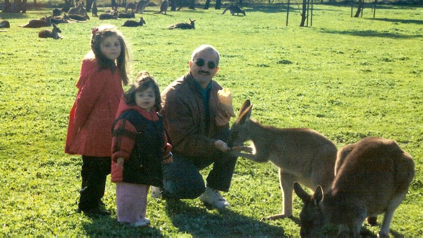 Two young girls stand next to their father who is crouching and feeding a kangaroo.
