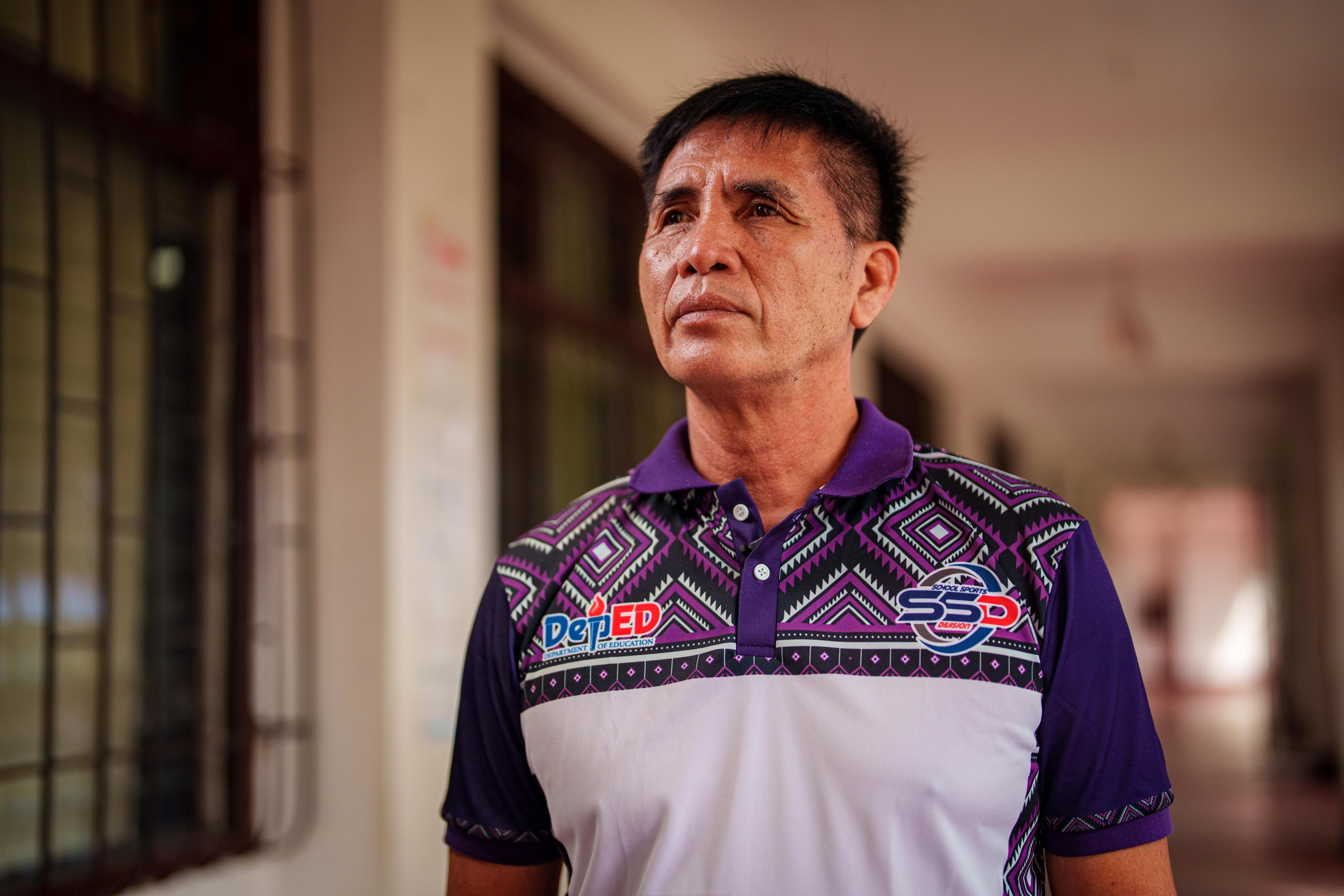 A middle-aged Filipino man wears a purple and white polo shirt standing in the hallway of classrooms.