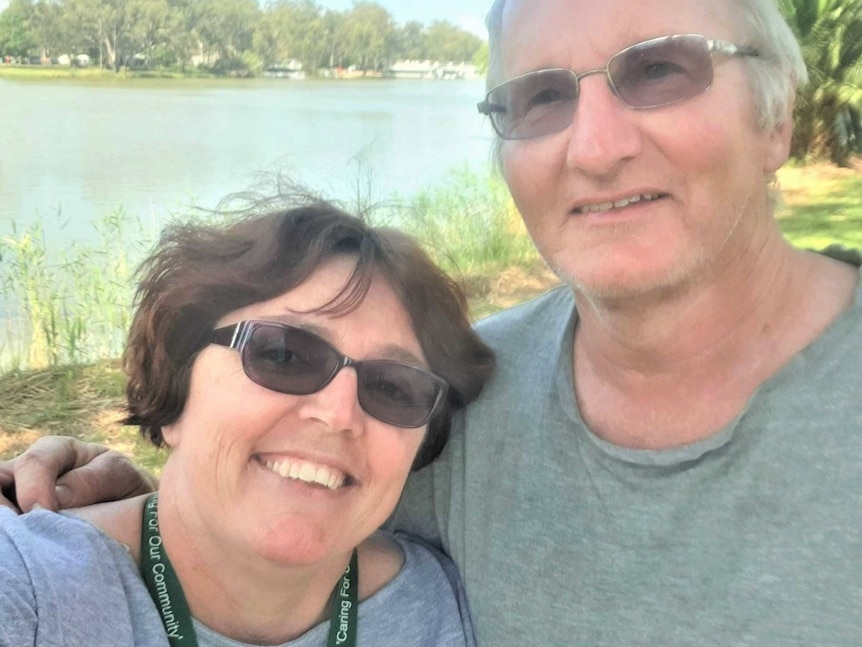 A man and woman with arms around each other grin and take a selfie, standing by the riverside.