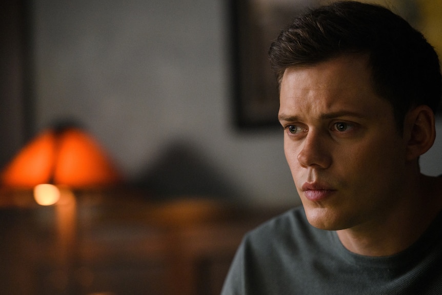 A young white man wearing a grey tshirt looks worried, a blurry lamp in the background