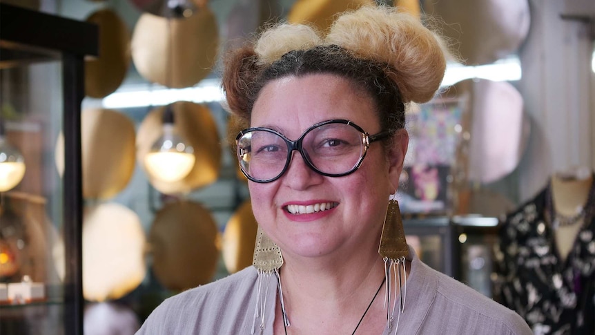 Ms Campbell, wearing large round glasses and large earrings, smiles while standing in her shop.