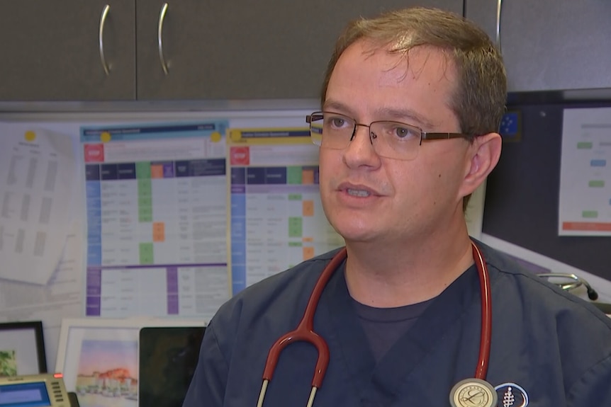 A man in blue medical scrubs with a stethoscope around his neck sitting in an office.