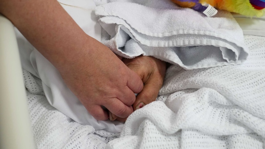 An elderly hand sits on white blankets, held by another hand from outside the bed.