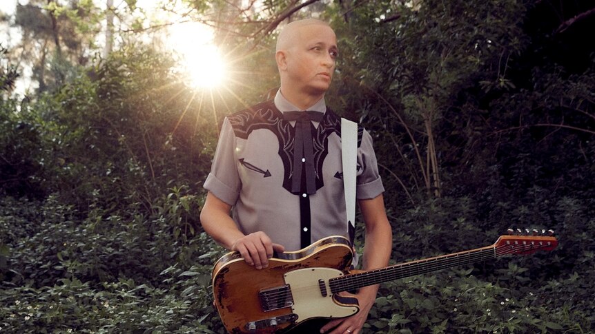 Mark Lizotte, AKA Diesel, standing in a forest with a guitar.
