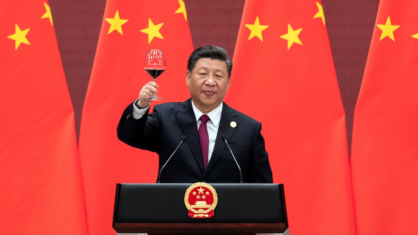 Xi Jinping raises his glass and proposes a toast while standing behind a lecturn in front of Chinese flags.